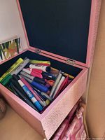 Open pink textured storage box filled with assorted markers, pens, and highlighters in various colors and brands.