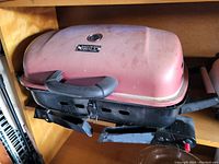 Photo shows a rectangular portable BBQ grill with a pink lid and black base, a handle on top of the lid and side handles, with vent holes on the front.