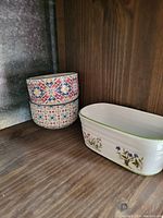 Three ceramic bowls on wooden shelf: two round multicolored patterned bowls stacked and one rectangular white floral bowl.