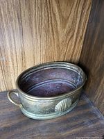 Oval brass bowl with a single handle and embossed shell pattern on the side, placed on wooden surface with wood background.