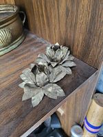 Photo showing two brass flower sculptures placed on a wooden shelf, showing their layered petals and leaves with natural patina.