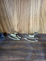 Two brass rocking horse figurines displayed on a wooden surface with visible grain and tone variations.