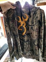 Camouflage hoodie sweater with orange Browning logo on chest, shown hanging and held by hand.