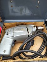 Close-up of a silver electric drill inside a metal case showing the drill's body, trigger, ventilation, and coiled black power cord.