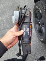 Black & Decker electric circular saw held in hand next to a tire on asphalt. Visible label shows 10 amps, 120V, 100mm blade size.