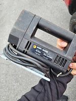 Black & Decker electric jig saw showing side with model and power specs, held in a hand, detail of body and power cord.