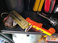 Fishing gear displayed on a shelf including Berkley Trilene XL fishing line, metal fish measuring ruler, orange Rapala handheld fish scale, and black flashlight.