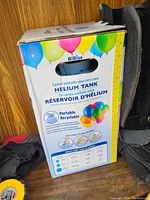 Photo of white and blue AirRise helium tank boxed with colorful balloons printed on the packaging. Visible instructional info and handle cutout.