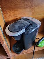 Front angled view of the Keurig coffee maker with silver lid and control buttons visible, showing dust accumulation on top.