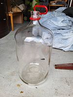 Photo showing a large clear glass jug with a wide round body and narrow neck, topped with a red plastic handle, placed on a concrete floor in a garage or storage area.