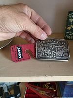 Hand holding embossed silver Levi's belt buckle next to red leather Levi's holder on wooden surface