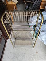 Gold-tone metal rack with three tiers and vertical wire dividers to hold records or items upright, shown standing on a concrete floor surrounded by other furniture and items.