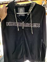 Front view of black zip-up hoodie sweater with Nickelback logo embroidered on chest and patterned inner hood lining.