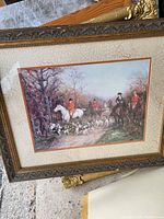 Full frontal view of the framed fox hunt print hanging or placed leaning on a textured surface, showing riders in red coats on horses with hunting dogs below, in an ornate carved frame.