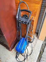Full view of the Simoniz electric pressure washer showing blue housing, black handle, hose, and wrapped power cord, placed beside furniture on concrete floor.