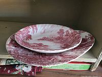 Two Ridgway Ironstone pieces stacked showing detailed red hand-engraved pastoral scenes on a white ceramic base, including houses, river scenes, and trees.