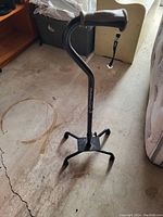 Photo showing a black walking cane with a curved handle and a four-pronged base standing upright on a concrete floor. The brand label is partly visible on the cane's shaft.