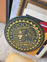 Round wooden wall sign with Waupoos Draft Cider branding, detailed logo and text, displayed leaning against other frames and signs.