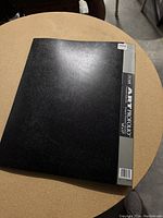 Closed black art portfolio showing the front cover and spine label with product details.