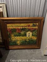 Photo of a framed landscape painting showing a small white house amidst colorful fields of vegetation with a multi-layered wooden frame against a gray corrugated metal backdrop.