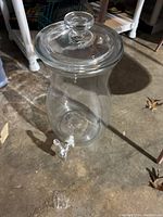 Photo showing clear glass beverage dispenser with rounded lid and white plastic spout on bottom side.