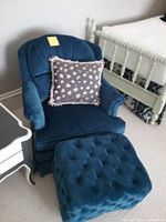 Blue velvet upholstered armchair with matching tufted ottoman in a room setting. Chair includes decorative heart-patterned pillow. Ottoman shows tufted buttons on top. One ottoman leg is damaged as noted.