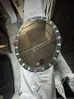 Top-down photo of an oval mirror with a decorative floral frame in various colors including purple, blue and pink. Mirror surface reflects ceiling and light.
