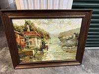 Framed oil painting showing waterfront village scene with houses, boats and trees in impressionistic style.