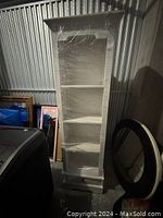 Full view of the tall white wooden bookshelf with three shelves, wrapped in protective plastic, stored in a dimly lit storage area.
