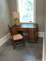 Photo showing a wooden desk with multiple drawers and leather inlay top alongside a wooden chair with woven seat situated in a room corner by a window.
