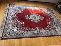 Full view of red area rug with cream border and floral medallion design laid flat on hardwood floor, fringe visible.