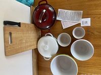 Top-down view of large red enamel pot with lid, wooden cutting board, white casserole dish with lid, and several small white ceramic bowls/cups.