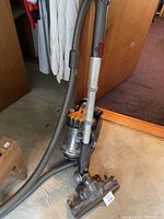 Photo of Dyson vacuum cleaner standing upright showing grey hose and orange cyclone head with clear dust container and metal wand.