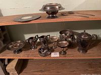 Wide view showing many silverplate tableware items arranged on wooden shelving including bowls, teapot, basket with tongs, warmer stand, and spoons.