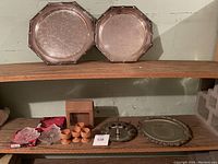 Shelf holding two large silver-tone serving trays on top shelf and assorted dishes and decor items on bottom shelf including wooden egg cups, silver-tone serving tray, crystal dishes, and ornate silver plate.