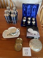 Photo shows six shiny silver egg cups in a blue velvet lined case, a black metal rack with six stainless steel condiment containers, a floral patterned cup and saucer with a matching dish, two round engraved metal coasters, and a boxed set of round silver-tone napkin holders on a wooden surface.