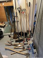 Wide shot showing a large number of garden tools leaning against a door and scattered on the floor, including shovels, rakes, a broom, pruning shears, a hoe, and other implements.
