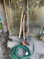 Photo showing a variety of garden tools including long-handled shovel, spade, hoe, pitchfork, rake, and a coiled green garden hose with a yellow and black attached spray nozzle.