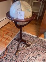 Photo showing a traditional nautical-style globe mounted on a wooden tripod stand with a round meridian, placed on a hardwood floor with a rug beneath.