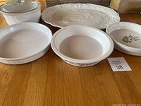 Top-down view of five dishes arranged on wooden surface: white ceramic oval platter with embossed turkey, round casserole with glass lid, two deep white bowls, and a small floral oval bowl.