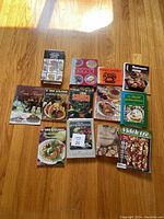 Photo showing 12 Hungarian cookbooks arranged on a wooden floor with various colorful covers depicting traditional Hungarian dishes and themes.