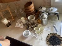 Photo showing a variety of kitchen items including glass jars, wooden bowls, pots, mugs, mortar and pestle, small cups, and egg cups on a countertop.