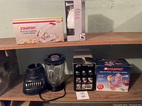 Photo of kitchen supplies on wooden shelves including boxed Starfrit potato chipper, electric blender, coffee grinders, and boxed Starfrit apple peeler.
