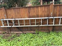 Folded aluminum ladder leaning horizontally on a wooden fence, with greenery below and a black hose or cable on it.