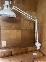 White adjustable desk lamp mounted on a wooden desk against a wood-paneled wall. Lamp has multi-joint arm and conical ventilated shade, secured with a clamp mount.