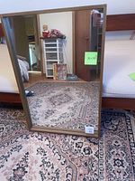 Front view of a large rectangular mirror with wooden frame, shown standing on a carpeted floor reflecting part of a room.