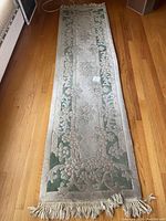 Full view of narrow carpet runner laid on wooden floor showing floral design in green and beige colors, with fringes at ends.
