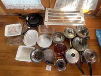 Overview of all pots, pans, baking dishes, lids, and other kitchenware laid on wooden floor near window.