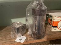 Retro coffee maker and matching cup, both wrapped in protective plastic, placed on a shelf with an orange and white box nearby.