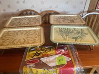 Four vintage metal TV trays on wooden table, two trays on each side, showing floral patterns and foldable metal legs. Plastic packaging with 'Lap Tray tables' label beneath trays.